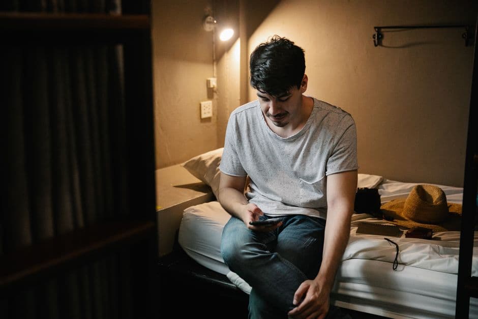 Young calm male in casual clothes sitting on comfortable bed and browsing mobile phone in hostel