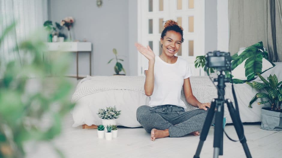 Smiling woman waves at camera while recording a vlog in a stylish bedroom setting.