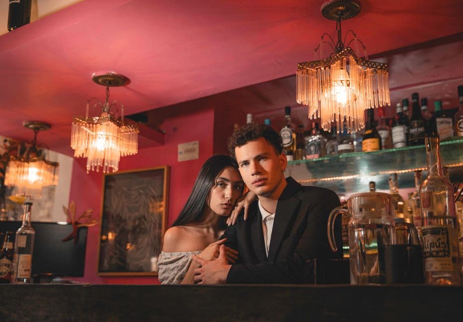 A stylish couple poses at a sophisticated bar, illuminated by warm, ambient lighting and chandeliers.