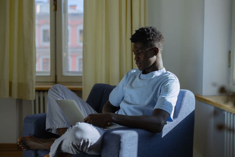 Man working remotely on a laptop while sitting on an armchair by the window in a cozy, sunlit room.