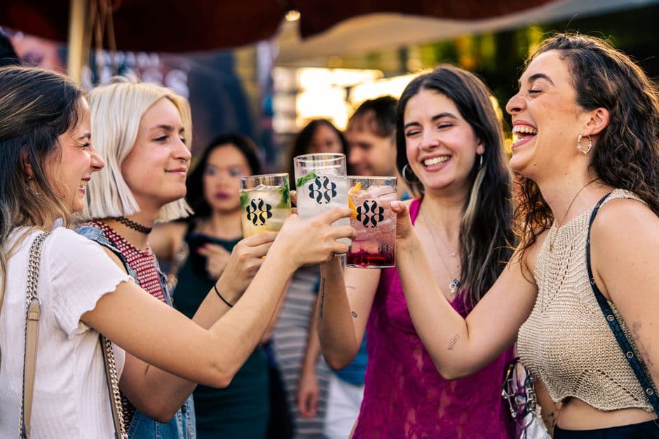 Young women laughing and clinking glasses in a lively outdoor setting, enjoying a festive atmosphere.