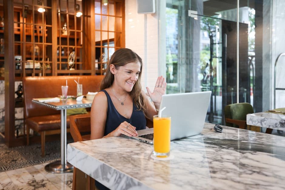 Smiling woman waving during an online meeting in a modern cafe with a laptop.
