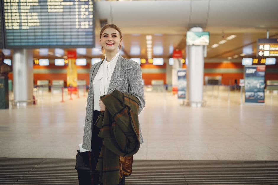Smiling female manager in elegant wear checking schedule of flights in airport while standing with suitcase and coat looking up