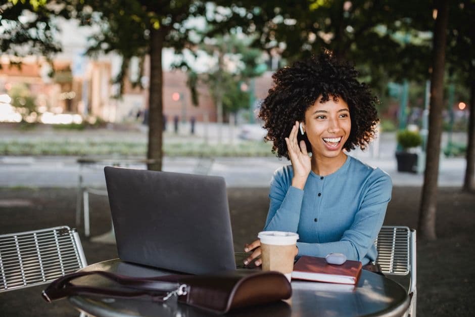 Smiling woman using a smartphone outdoors at a cafe with a laptop, enjoying a casual moment.