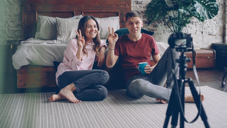 Happy couple sitting on floor recording a vlog in a cozy bedroom.